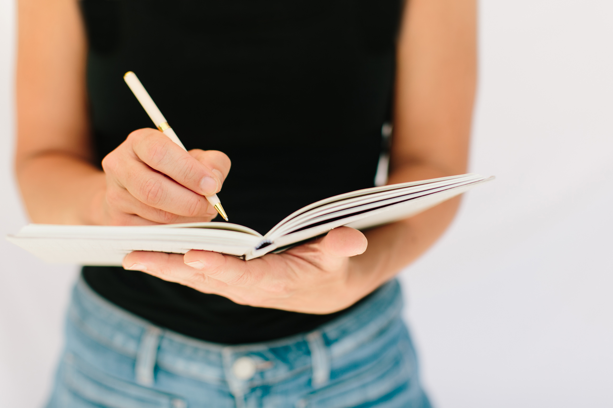 Image of a woman's torso, her hands holding a notebook in front of her and writing.
